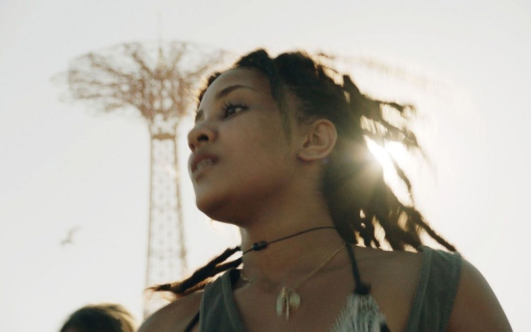 Filmstill aus TENDABERRY: Man sieht eine junge Frau, aus der Untersicht und leicht unscharf aufgenommen, an einem sonnigen Tag.