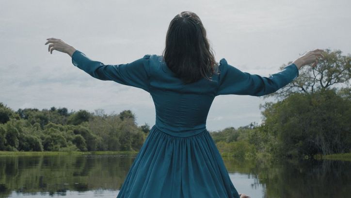 Film still from LUZ NOS TROPICOS. We see a woman in a blue dress standing in front of a lake from behind.