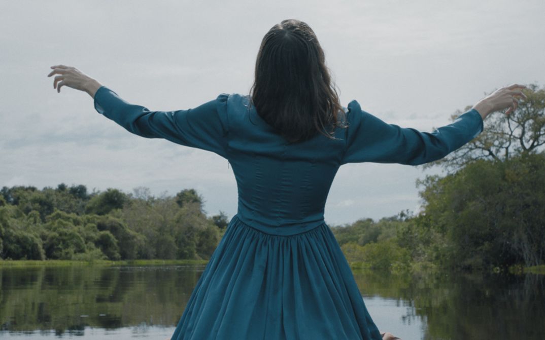 Film still from LUZ NOS TROPICOS. We see a woman in a blue dress standing in front of a lake from behind.