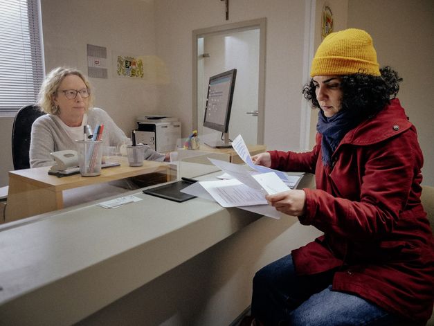 Filmstill aus "Shahid" von Narges Kalhor. Zu sehen ist eine sitzende Frau in einer roten Jacke, die Papiere liest. Ihr gegenüber sitzt eine Frau an einem Computer und schaut sie an. 