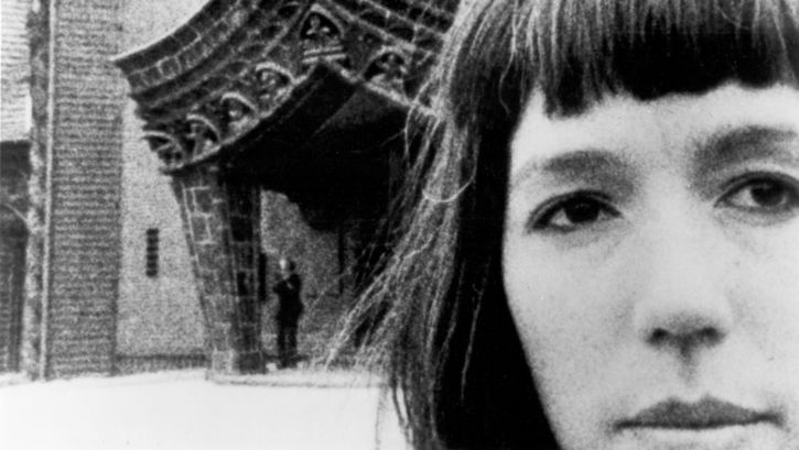 Filmstill aus JOURNEYS FROM BERLIN/1971: Ein Frauengesicht in Großaufnahme, dahinter ein auffällig gebautes Tor.