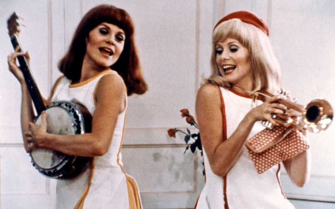 Film still from LES DEMOISELLES DE ROCHEFORT: Two young women in white summer dresses are dancing. One is holding a trumpet, the other is playing a banjo.