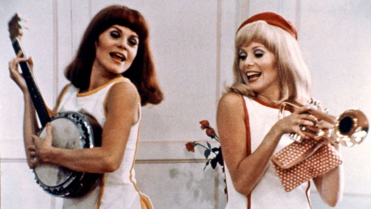 Film still from LES DEMOISELLES DE ROCHEFORT: Two young women in white summer dresses are dancing. One is holding a trumpet, the other is playing a banjo.