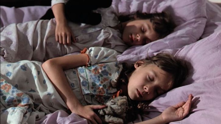 Film still from STAUNEN: two young girls sitting lying next to each other in bed, asleep. One is holding a cuddle toy, the sheets are light purple.