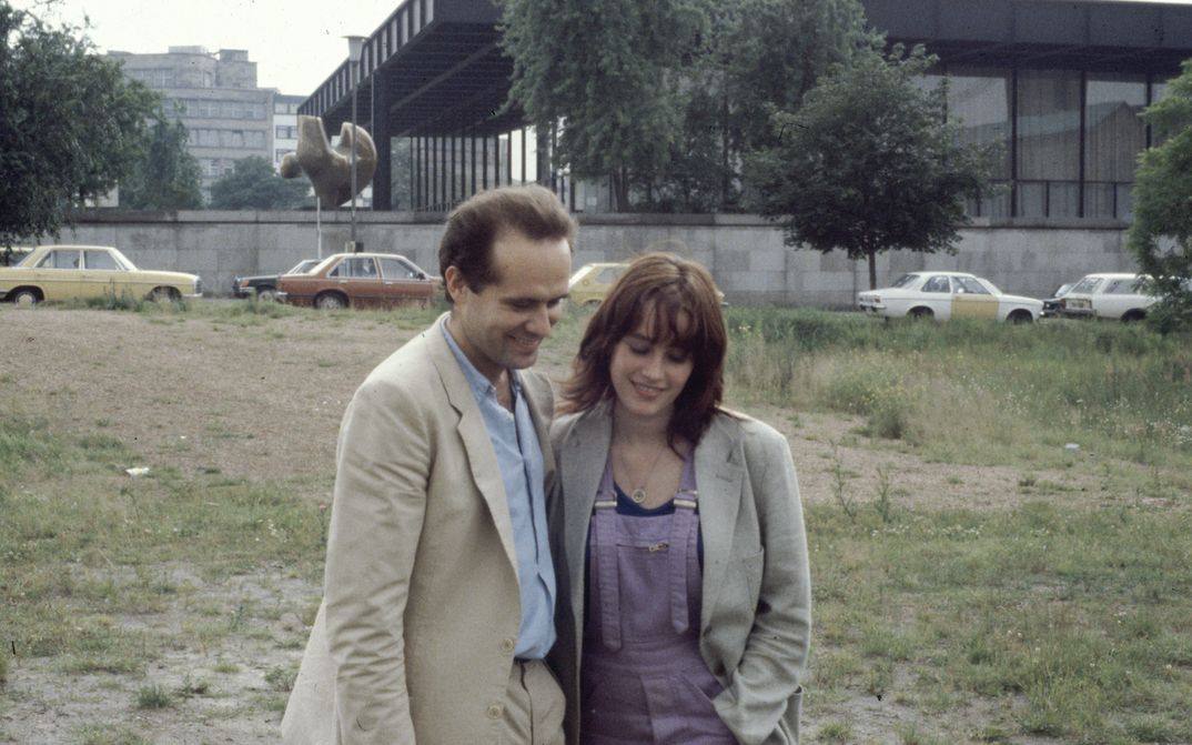 Filmstill aus BERLIN CHAMISSOPLATZ: Ein Mann und eine Frau stehen nah zusammen auf einer Brachfläche vor der Neuen Nationalgalerie in Berlin.