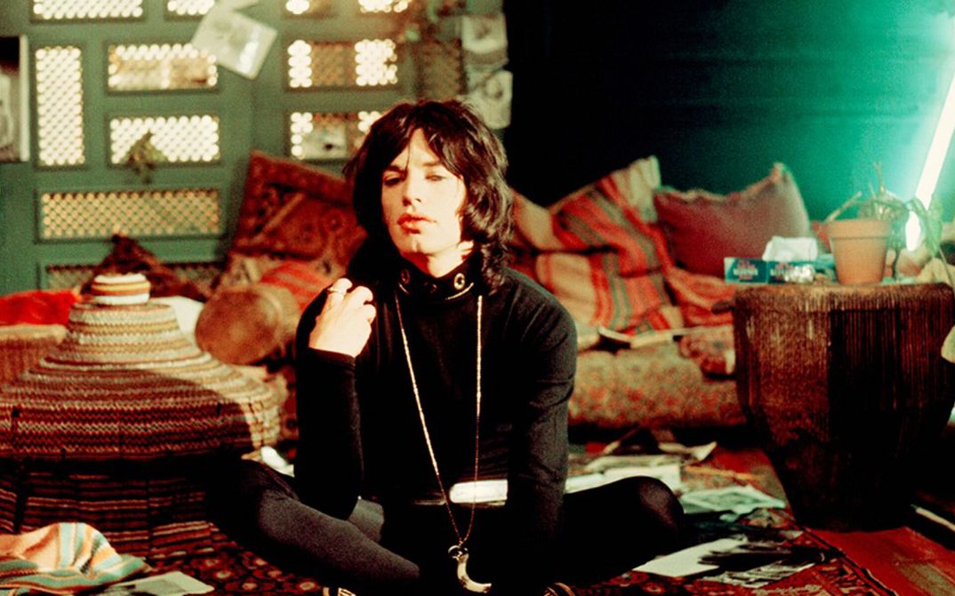 Film still from PERFORMANCE: Mick Jagger, dressed in black, is sitting cross-legged on the floor of a room furnished with colorful furniture and rugs.