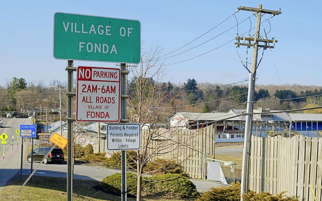 Filmstill aus "Henry Fonda for President" von Alexander Horwath. Zu sehen ist das Ortseingangsschild von Fonda. Im Hintergrund sind eine Straße und Häuser.