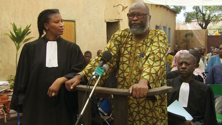 Film still from BAMAKO: An improvised courtroom scene in the open air. In the foreground a woman in a judge
