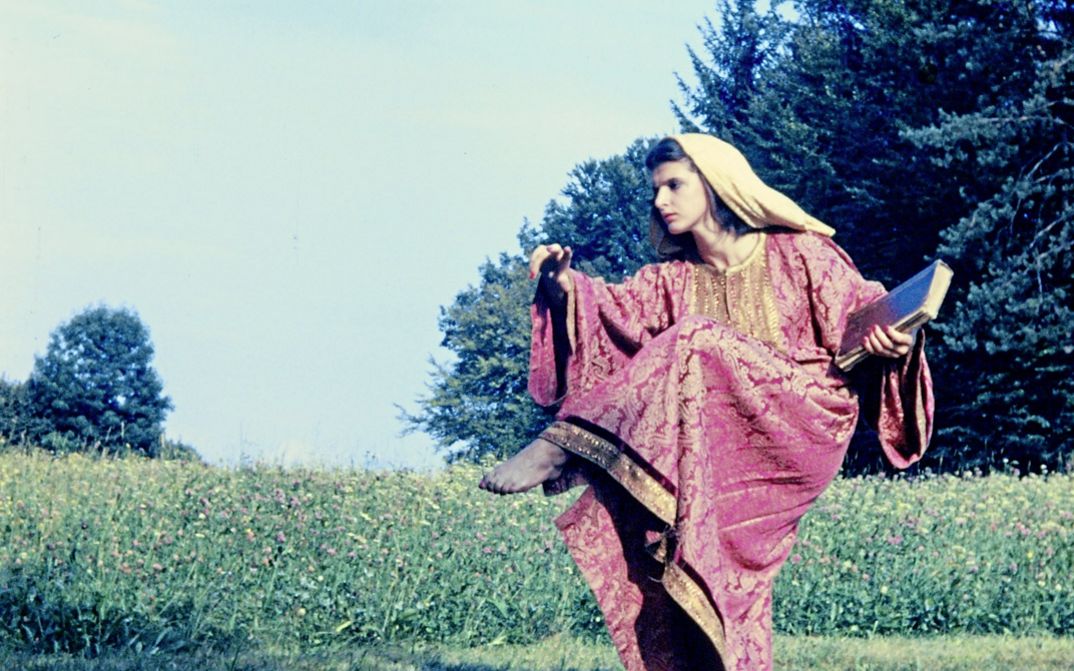 Filmstill from "Baroque Statues" by Maria Lassnig. It shows a woman in a red robe and with a scarf on her head. She raises her leg. A wildflower meadow can be seen in the background. 