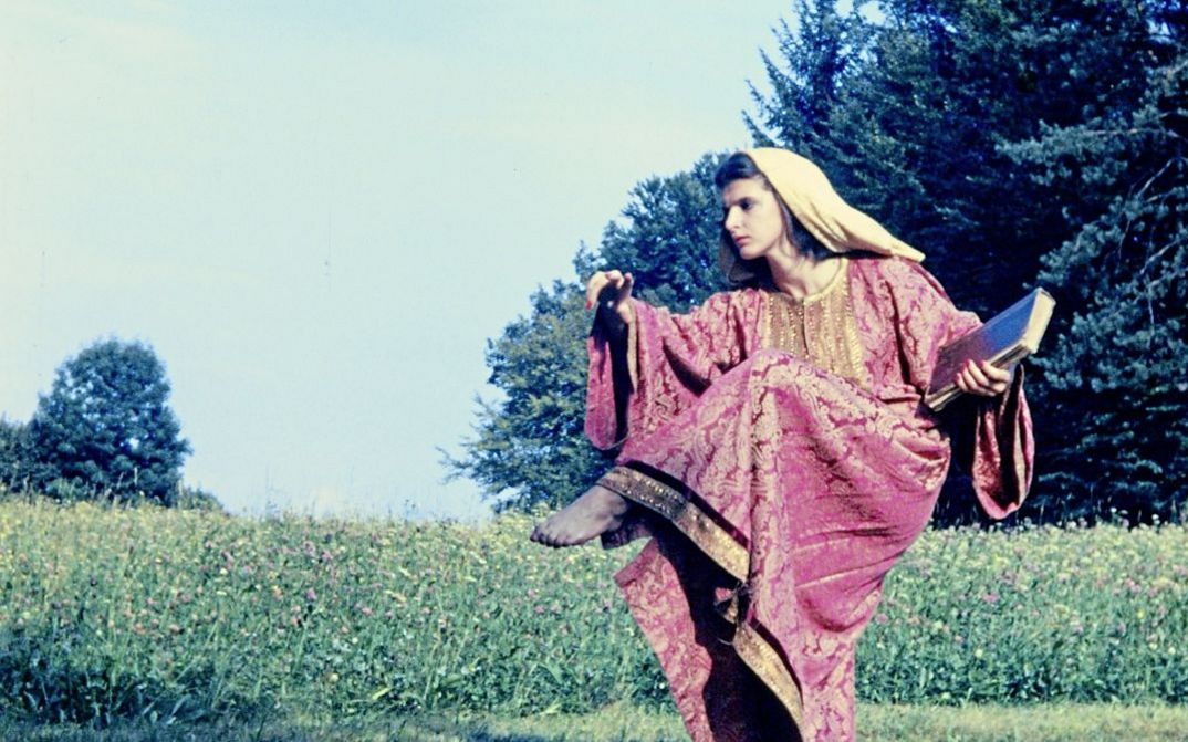 Filmstill aus "Baroque Statues" von Maria Lassnig. Zu sehen ist eine Frau in einem roten Gewand und mit einem Tuch auf dem Kopf. Sie hebt ihr Bein hoch. Im Hintergrund ist eine Wildblumenwiese.