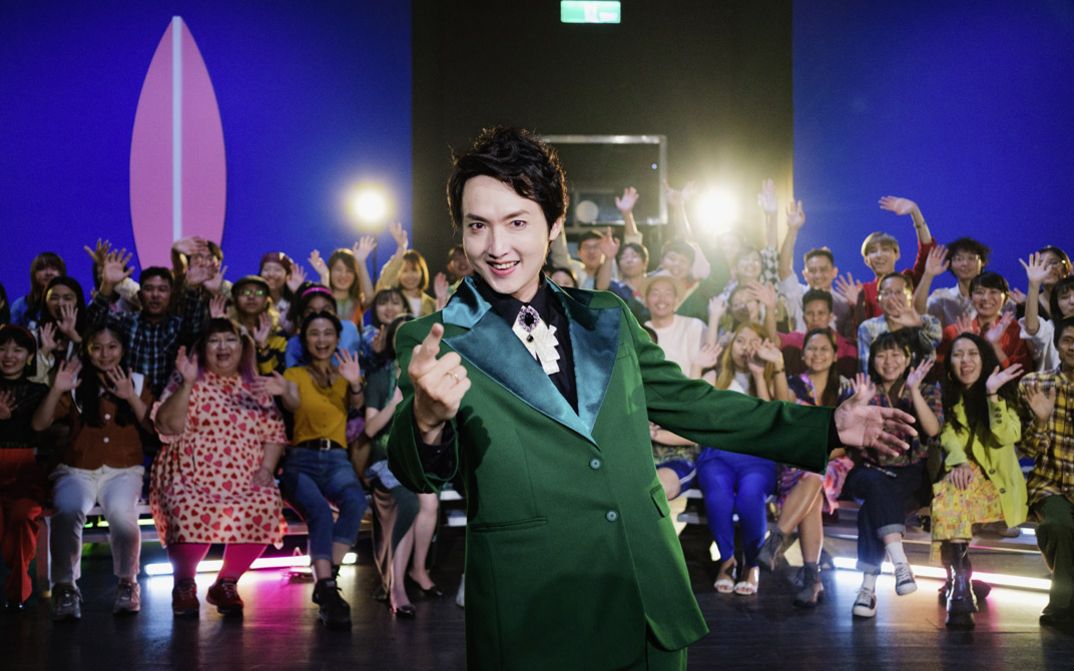A man in a green suit is standing in a television studio. He looks into the camera and points forwards with his hand.