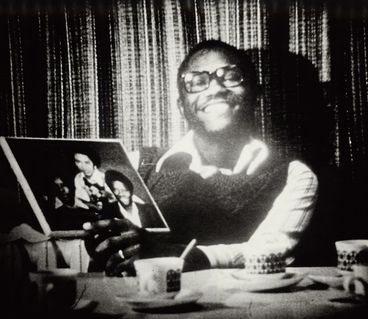 Black and white image of a young man sitting at a table smiling and showing a family photo to the camera.