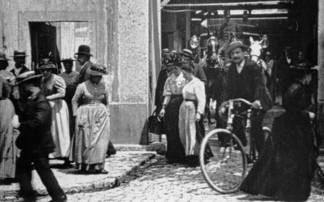 Filmstill from ARBEITER VERLASSEN DIE FABRIK: A street scene; many people in old clothes, some with hats. A man on a bicycle. A carriage in the background.