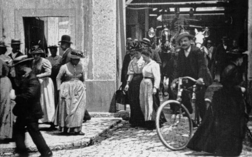Filmstill aus ARBEITER VERLASSEN DIE FABRIK: Eine Straßenszene; viele Menschen in alten Kleidern, teils mit Hüten. Ein Mann auf dem Fahrrad. Im Hintergrund eine Kutsche.