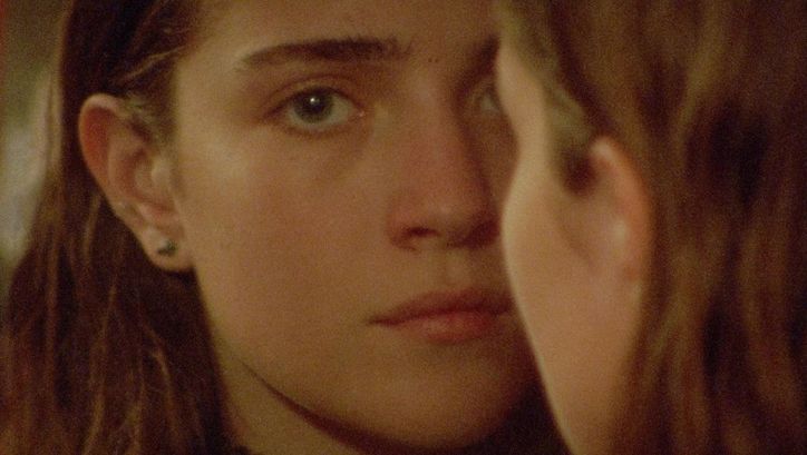 Film still from DIESES ZIMMER KANN MAN NICHT ESSEN: A young girl with long hair looks into the camera. Another person with long hair can be seen from behind, half covering her face.