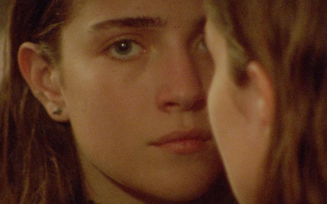 Filmstill aus DIESES ZIMMER KANN MAN NICHT ESSEN: Ein junges Mädchen mit langen Haaren blickt in die Kamera. Eine andere Persoon mit langen Haaren ist von hinten zu sehen und verdeckt halb ihr Gesicht.
