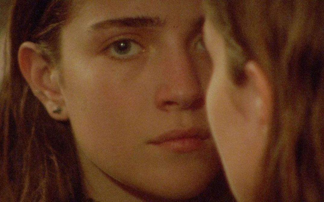 Film still from DIESES ZIMMER KANN MAN NICHT ESSEN: A young girl with long hair looks into the camera. Another person with long hair can be seen from behind, half covering her face.
