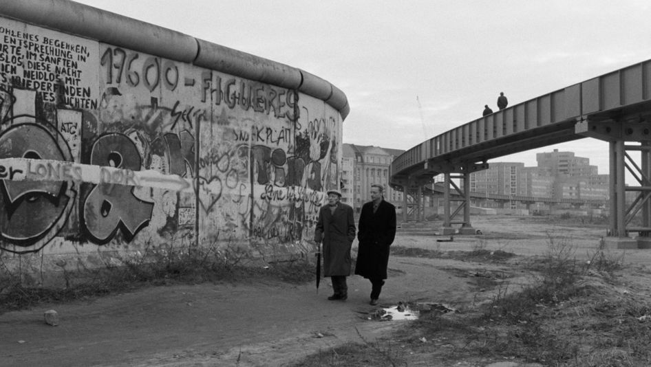 Film still from DER HIMMEL ÜBER BERLIN: Two men walk across a wasteland, the painted Berlin Wall to their left.