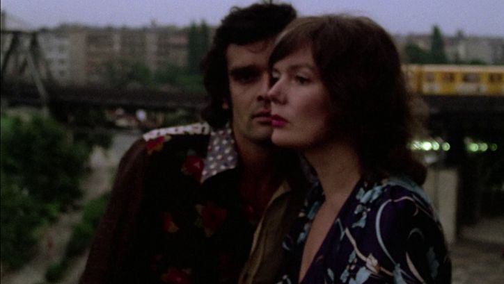 Filmstill aus ERZÄHLEN. Die Gesichter eines Mannes und einer Frau halb übereinander. Im Hintergrund die Berliner Hochbahn.