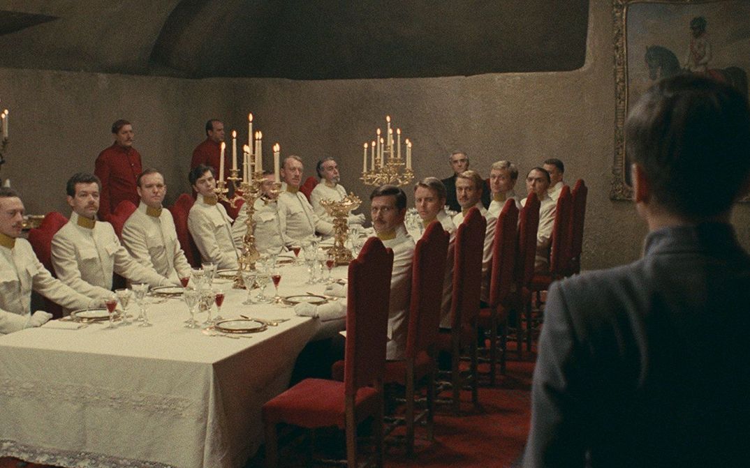 Filmstill aus Il deserto dei tartari: An einer langen, edel gedeckten Tafel sitzen Offiziere in weißen Uniformen. Alle blicken in eine Ecke, in der ein jüngerer Mann steht.