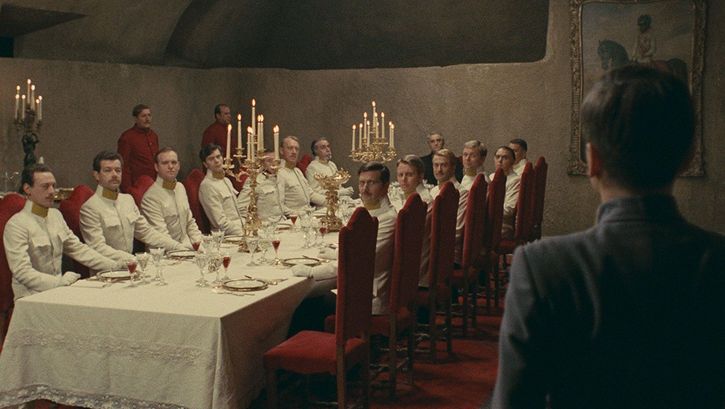 Filmstill aus Il deserto dei tartari: An einer langen, edel gedeckten Tafel sitzen Offiziere in weißen Uniformen. Alle blicken in eine Ecke, in der ein jüngerer Mann steht.