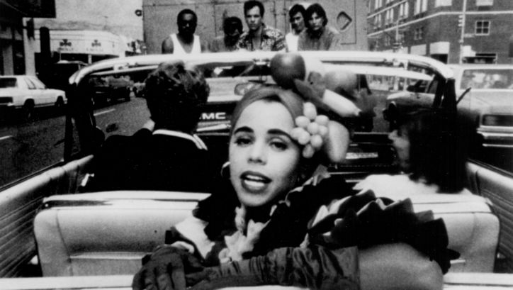 Film still from PRIVILEGE: A woman turns to the viewer in the back seat of a convertible.