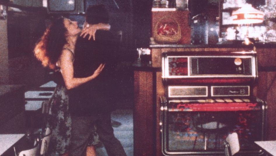 Film still from TOUTE UNE NUIT: In an otherwise empty bar, a woman and a man are dancing, holding each other tightly. A jukebox stands next to them.
