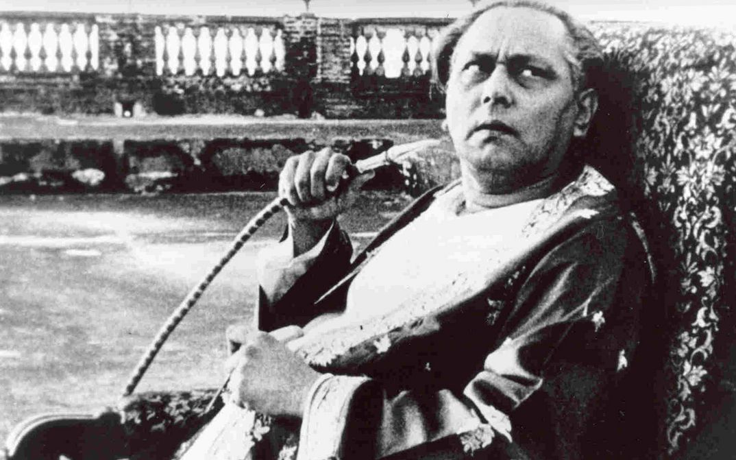Filmstill aus JALSAGHAR: Ein älterer Mann sitz auf einem Sessel, blickt skeptisch nach oben und hält einen Shisha-Schlauch in der Hand.