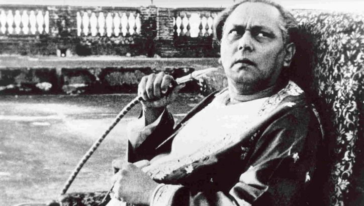Filmstill aus JALSAGHAR: Ein älterer Mann sitz auf einem Sessel, blickt skeptisch nach oben und hält einen Shisha-Schlauch in der Hand.