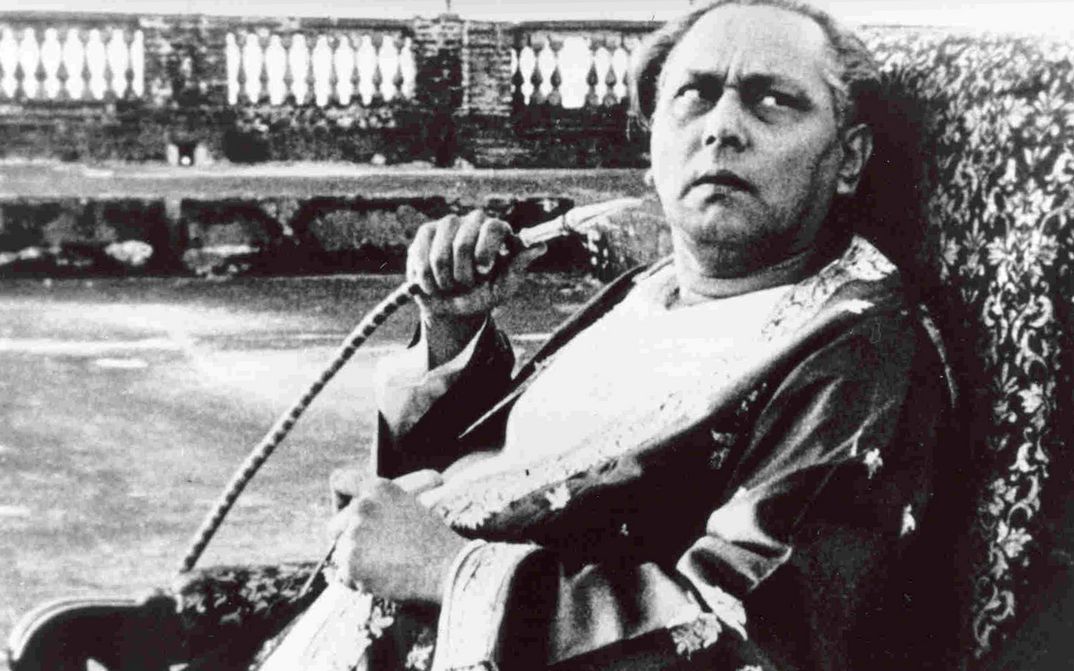 Filmstill aus JALSAGHAR: Ein älterer Mann sitz auf einem Sessel, blickt skeptisch nach oben und hält einen Shisha-Schlauch in der Hand.