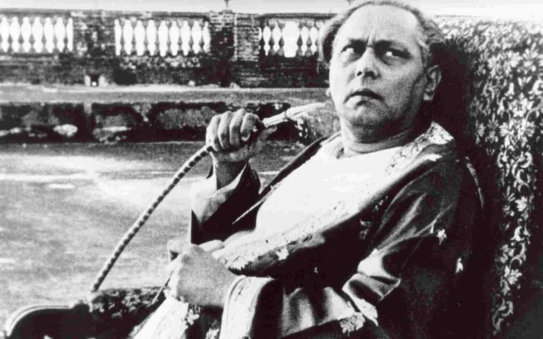 Filmstill aus JALSAGHAR: Ein älterer Mann sitz auf einem Sessel, blickt skeptisch nach oben und hält einen Shisha-Schlauch in der Hand.