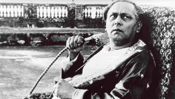 Filmstill from JALSAGHAR: An older man sits on an armchair, looks up sceptically and holds a shisha hose in his hand.