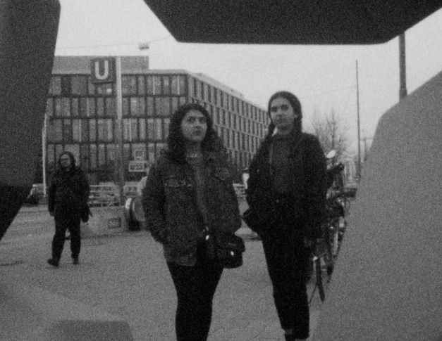 Filmstill aus „This Makes Me Want to Predict the Past“ von Cana Bilir-Meier. Zwei junge Frauen in einer Stadtlandschaft. Im Hintergrund ein modernes Gebäude. 