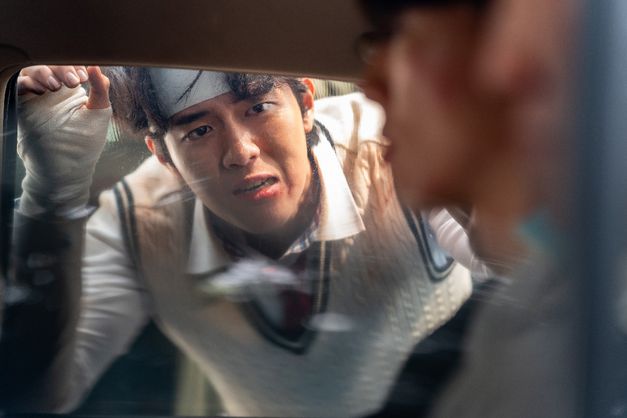 Eine Person mit bandagierter Hand beugt sich von außen an ein Autofenster und blickt zu einer Person im Inneren.