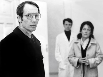 Black and white image of a man wearing glasses and a dark coat. In the background are a woman in a trench coat and a doctor in a lab coat with her hands in her pockets.