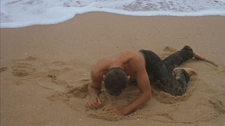 Filmstill aus ORÁCULO: Ein Mann mit nacktem Oberkörper gräbt sich an einem Strand in den Sand.