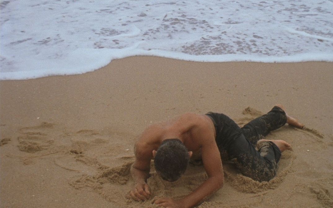 Filmstill aus ORÁCULO: Ein Mann mit nacktem Oberkörper gräbt sich an einem Strand in den Sand.