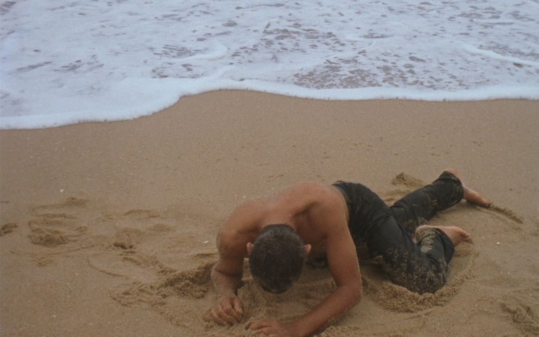Film still from ORÁCULO: A bare-chested man digs himself into the sand on a beach.