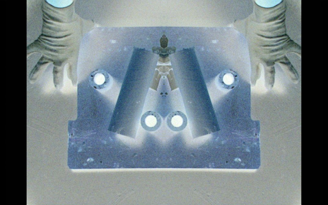 Film still from Crystal Z Campbell’s „Revolver“. Top view onto a vertically mirrored object with abstract forms on it, held by a hand on each side.