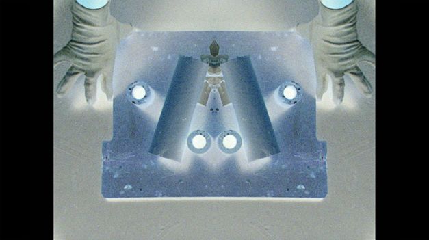 Film still from Crystal Z Campbell’s „Revolver“. Top view onto a vertically mirrored object with abstract forms on it, held by a hand on each side.