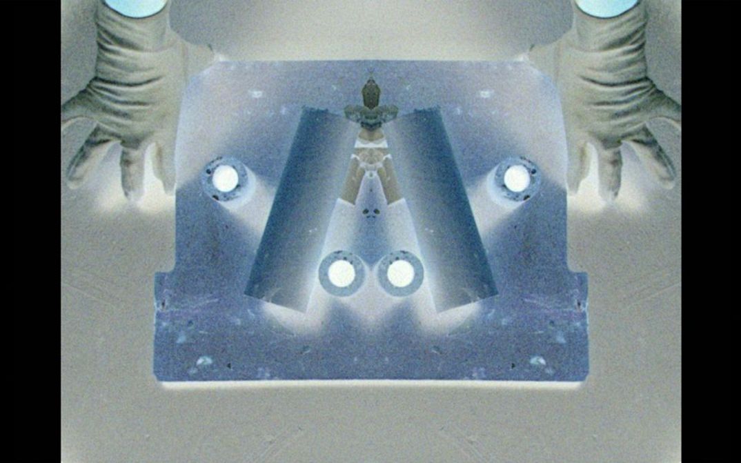 Filmstill aus dem Film „Revolver“ von Crystal Z Campbell. Aufsicht auf ein in der Mitte gespiegeltes Objekt mit abstrakten Formen darauf, welches auf beiden Seiten von jeweils einer Hand gehalten wird.