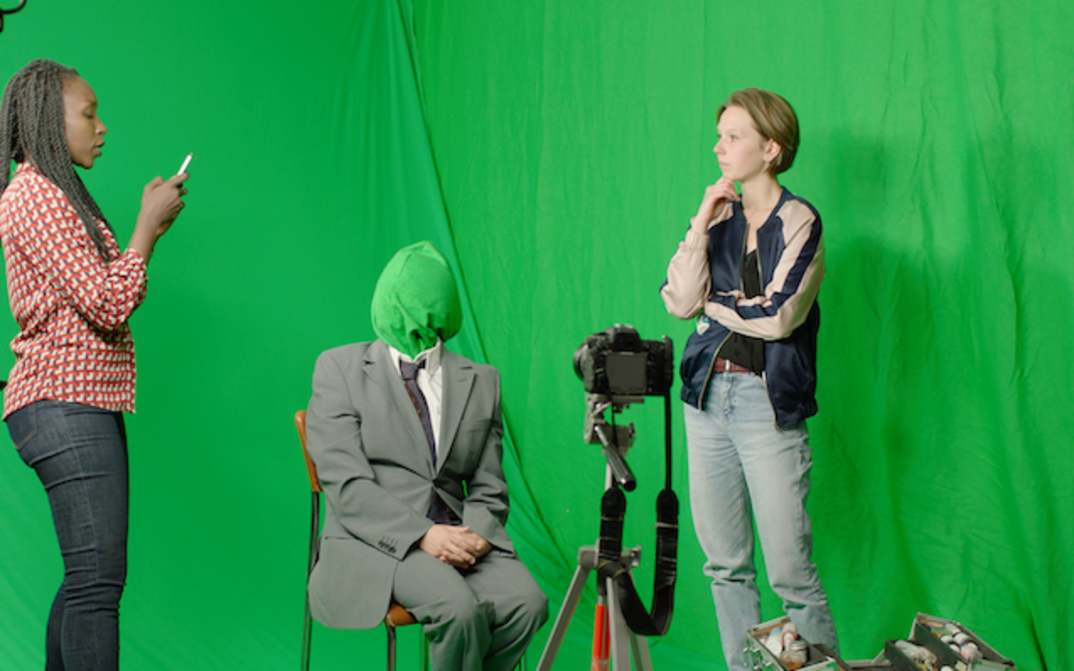 Film still from TÉLÉ RÉALITÉ: A person wearing a suit and a green hood pulled over their head is sitting in front of a camera. Two women are standing to the left and right of them. The background is covered with a green cloth. 