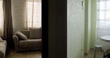 Still from the film „Llamadas desde Moscú“ by Luís Alejandro Yero. The interior of a flat. In the left room there are two sofas and windows with curtains. In the right room, a table with a stool and a window with curtains.