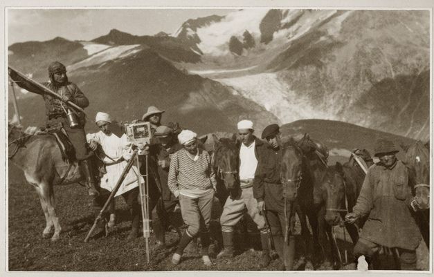 Filmstill aus "Deda-Shvili an rame ar aris arasodes bolomde bneli" von Lana Gogoberidze. Zu sehen ist eine Reihe von Personen und Pferden in den Bergen. Auf der linken Seite ist eine Kamera. 