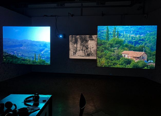 Film Still aus „Industries of Denial, Stage 10: From Musa Dagh to Port Saïd“ von Angela Melitopoulos und Kerstin Schroedinger. Eine dunkle Ausstellungsinstallation zeigt drei nebeneinander angeordnete Projektionen: links und rechts farbintensive Landschaftsbilder mit grünen Hügeln und Häusern, in der Mitte eine kleinere, schwarz-weiße historische Fotografie.  