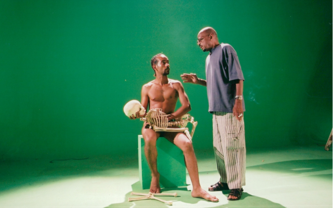Filmstill aus OF DUST AND RUBIES: Vor einem grünen Hintergrund sind zwei Männer zu sehen. Einer der beiden ist nackt und hält ein Skelett vor sich.