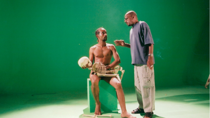 Film still from OF DUST AND RUBIES: Two men can be seen against a green background. One of them is naked and holding a skeleton in front of him. 