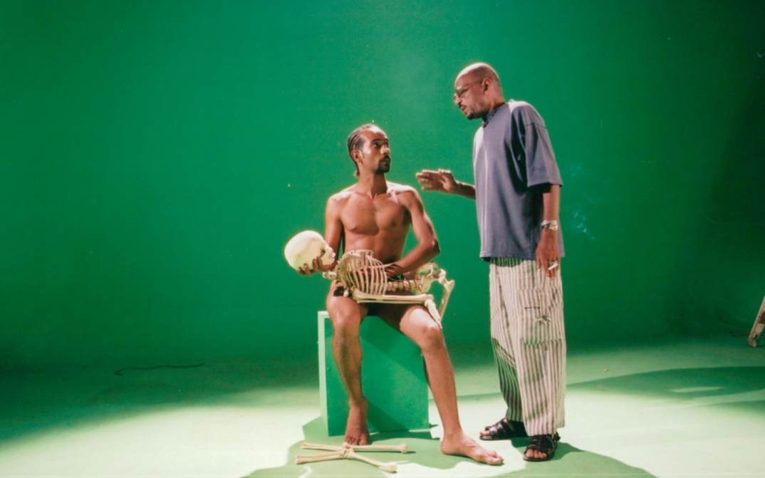 Filmstill aus OF DUST AND RUBIES: Vor einem grünen Hintergrund sind zwei Männer zu sehen. Einer der beiden ist nackt und hält ein Skelett vor sich. 