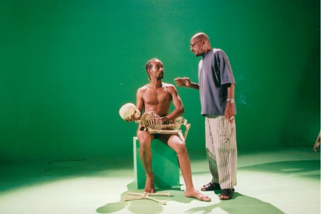 Filmstill aus OF DUST AND RUBIES: Vor einem grünen Hintergrund sind zwei Männer zu sehen. Einer der beiden ist nackt und hält ein Skelett vor sich. 