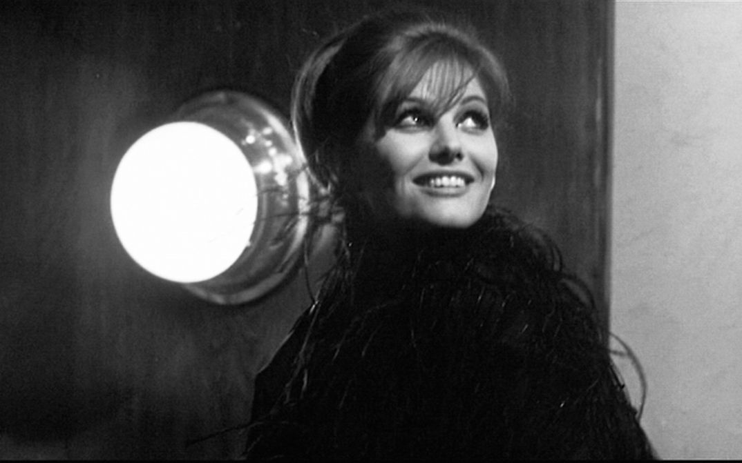 Film still from OTTO E MEZZO: A smiling young woman looks up at the sky, a bright lamp shines on the wall next to her.